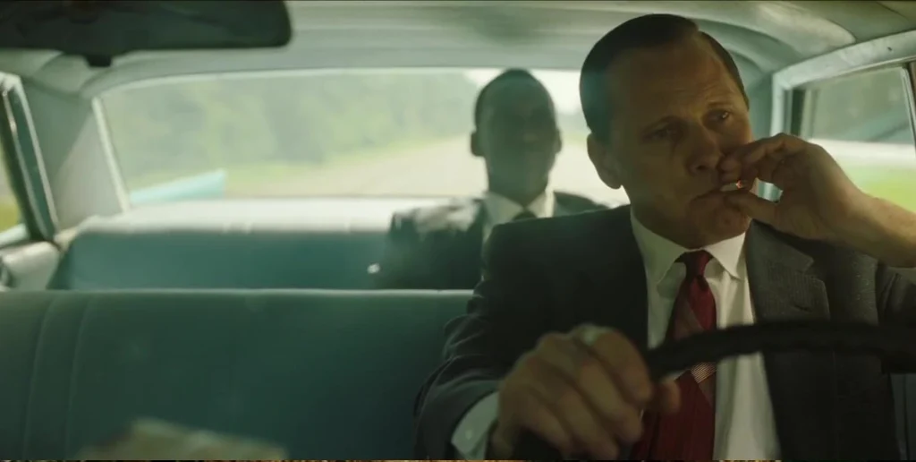 Green Book (2018): Sebuah Perjalanan Penuh Tawa dan Makna Dua pria berada di dalam mobil. Pria di depan mengemudi sambil merokok, mengenakan jas dan dasi merah. Pria di belakang duduk dengan santai. Jalan dan pemandangan hijau terlihat melalui jendela mobil.